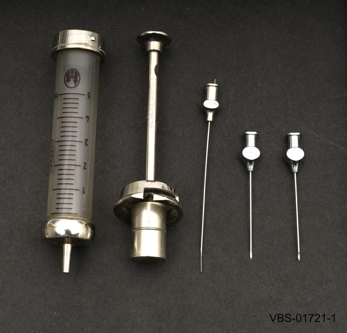 5ml sprøyte med beholder som ligger demontert i to deler, sammen med 3 sprøytespisser (nr. 12) og en stålsonde for å rengjøre nålen på innsiden.
Beholderen er en metallboks/etui i rusfritt stål med et stempel på lokket.