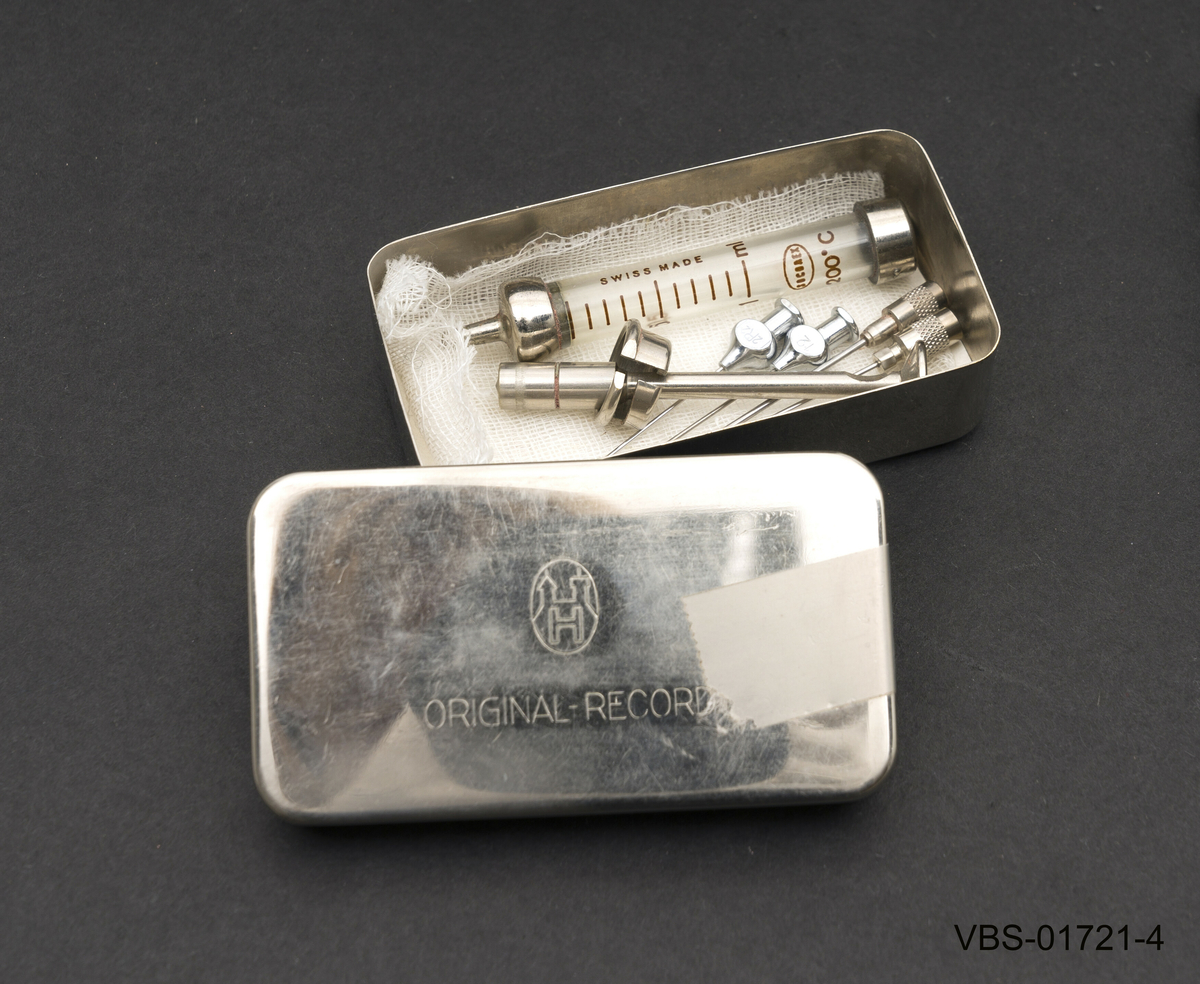 2 ml sprøyte med beholder som ligger demontert i to deler, sammen med 4stk. sprøytesipsser (nr. 12) med koniske hoder.
Beholderen er en metallboks i rusfritt stål og har et steril bomull gasbind på bunnen.