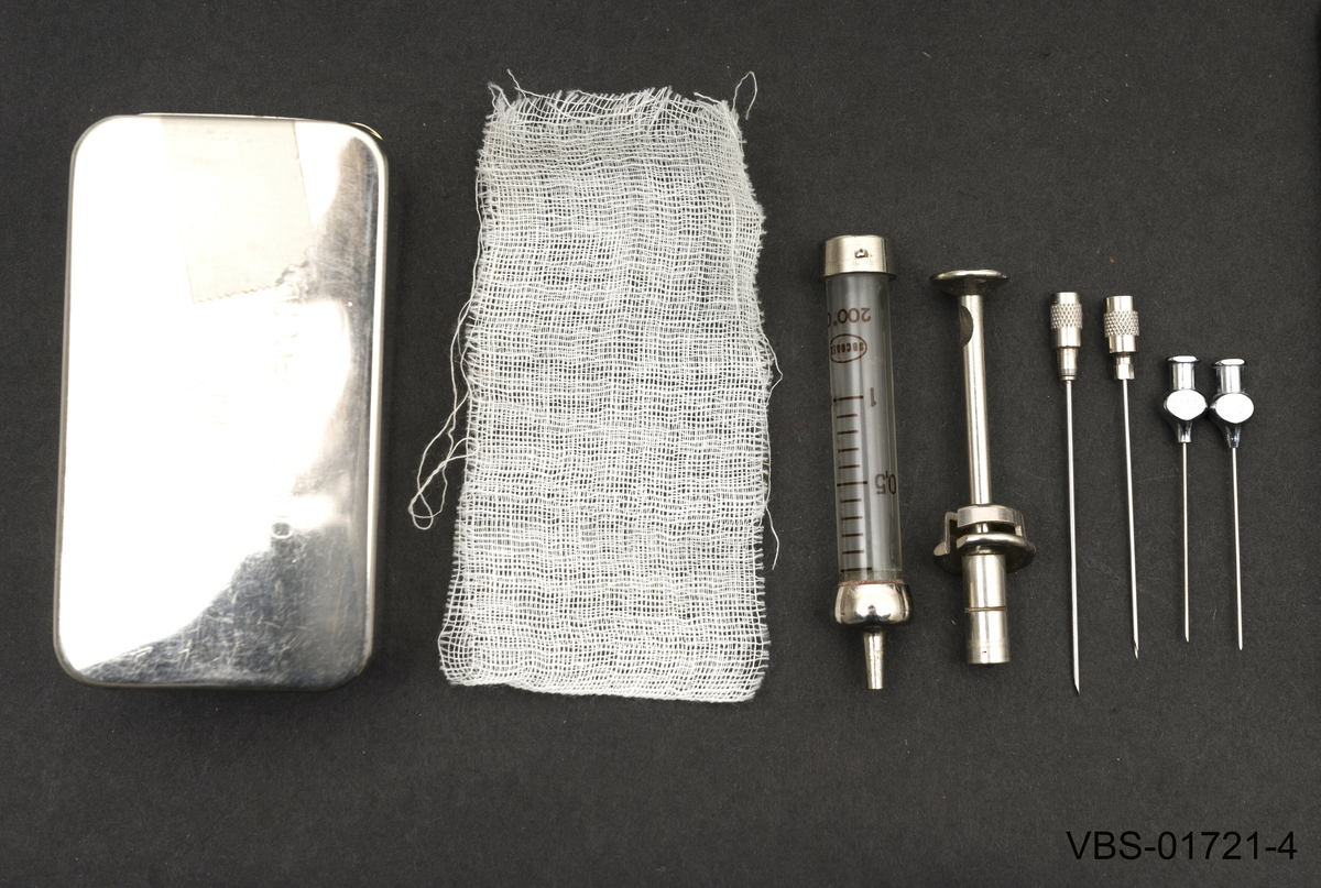 2 ml sprøyte med beholder som ligger demontert i to deler, sammen med 4stk. sprøytesipsser (nr. 12) med koniske hoder.
Beholderen er en metallboks i rusfritt stål og har et steril bomull gasbind på bunnen.