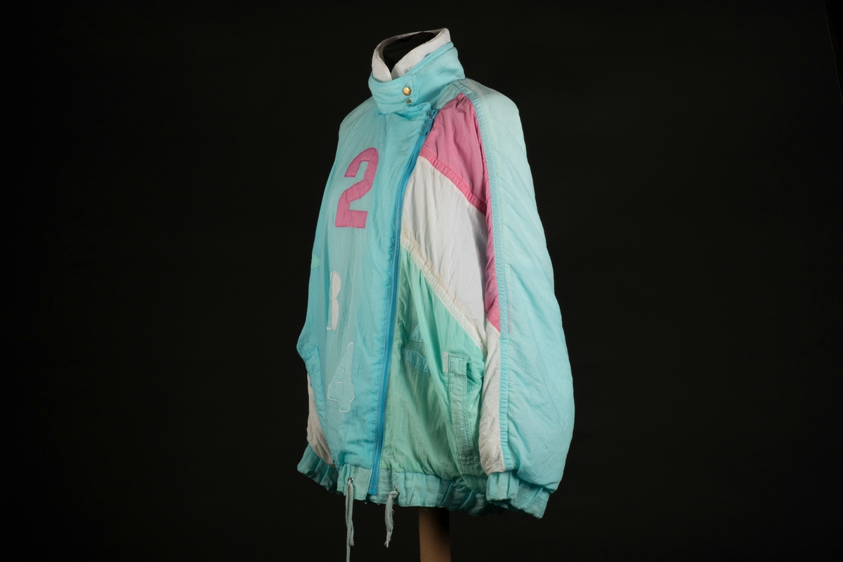 Skidjacka i pingvinmodell tillverkad av syntetmaterial, nylon/polyester.
Jackan i turkost, rosa, mintgrönt och vitt är sydd i så kallade pingvinmodell. Mönstret består av oregelbundna fält med de olika färgerna. På framsidan finns ditsydda applikationer med siffrorna 1 till 5 i olika färger. Den ljusblå dragkedjan är placerad något till vänster och diagonalt på framsidan. Den upprättstående kragen är försedd med två tryckknappar, och innanför finns ytterligare en krage. Det finns två fickor, försedda med dragkedjor. Jackan har insydda resårer nedtill samt i ärmsluten. Nedtill finns det även en dragsko med snodd.