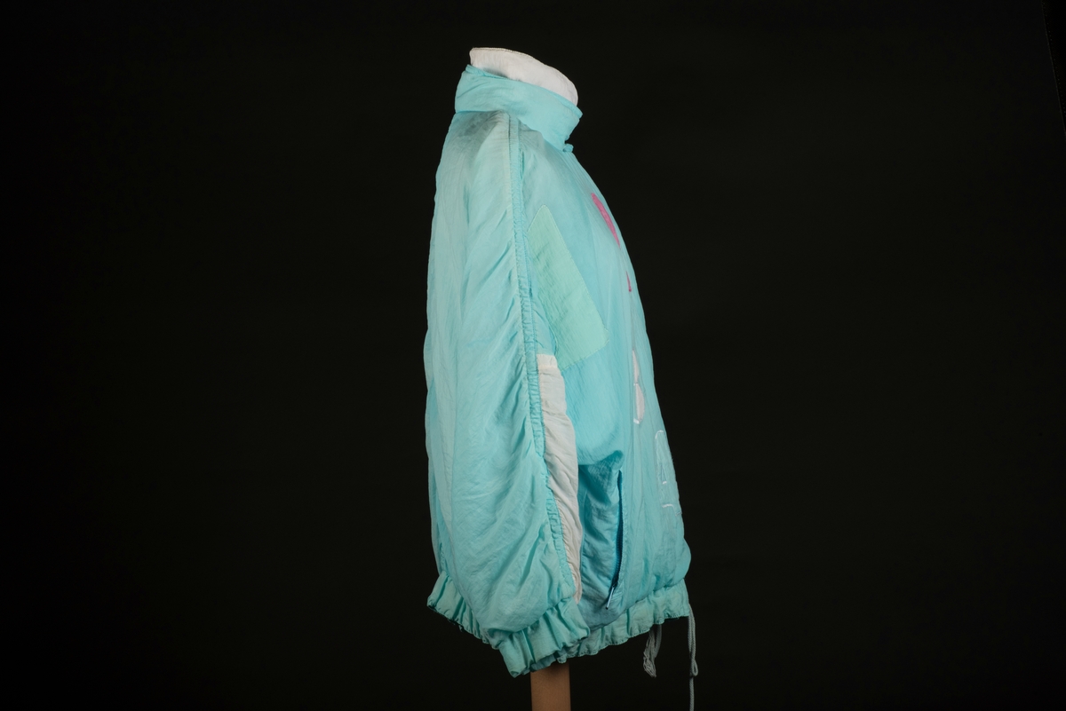 Skidjacka i pingvinmodell tillverkad av syntetmaterial, nylon/polyester.
Jackan i turkost, rosa, mintgrönt och vitt är sydd i så kallade pingvinmodell. Mönstret består av oregelbundna fält med de olika färgerna. På framsidan finns ditsydda applikationer med siffrorna 1 till 5 i olika färger. Den ljusblå dragkedjan är placerad något till vänster och diagonalt på framsidan. Den upprättstående kragen är försedd med två tryckknappar, och innanför finns ytterligare en krage. Det finns två fickor, försedda med dragkedjor. Jackan har insydda resårer nedtill samt i ärmsluten. Nedtill finns det även en dragsko med snodd.