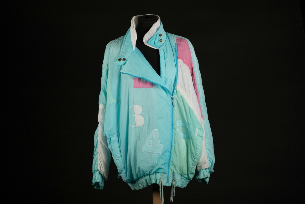 Skidjacka i pingvinmodell tillverkad av syntetmaterial, nylon/polyester.
Jackan i turkost, rosa, mintgrönt och vitt är sydd i så kallade pingvinmodell. Mönstret består av oregelbundna fält med de olika färgerna. På framsidan finns ditsydda applikationer med siffrorna 1 till 5 i olika färger. Den ljusblå dragkedjan är placerad något till vänster och diagonalt på framsidan. Den upprättstående kragen är försedd med två tryckknappar, och innanför finns ytterligare en krage. Det finns två fickor, försedda med dragkedjor. Jackan har insydda resårer nedtill samt i ärmsluten. Nedtill finns det även en dragsko med snodd.