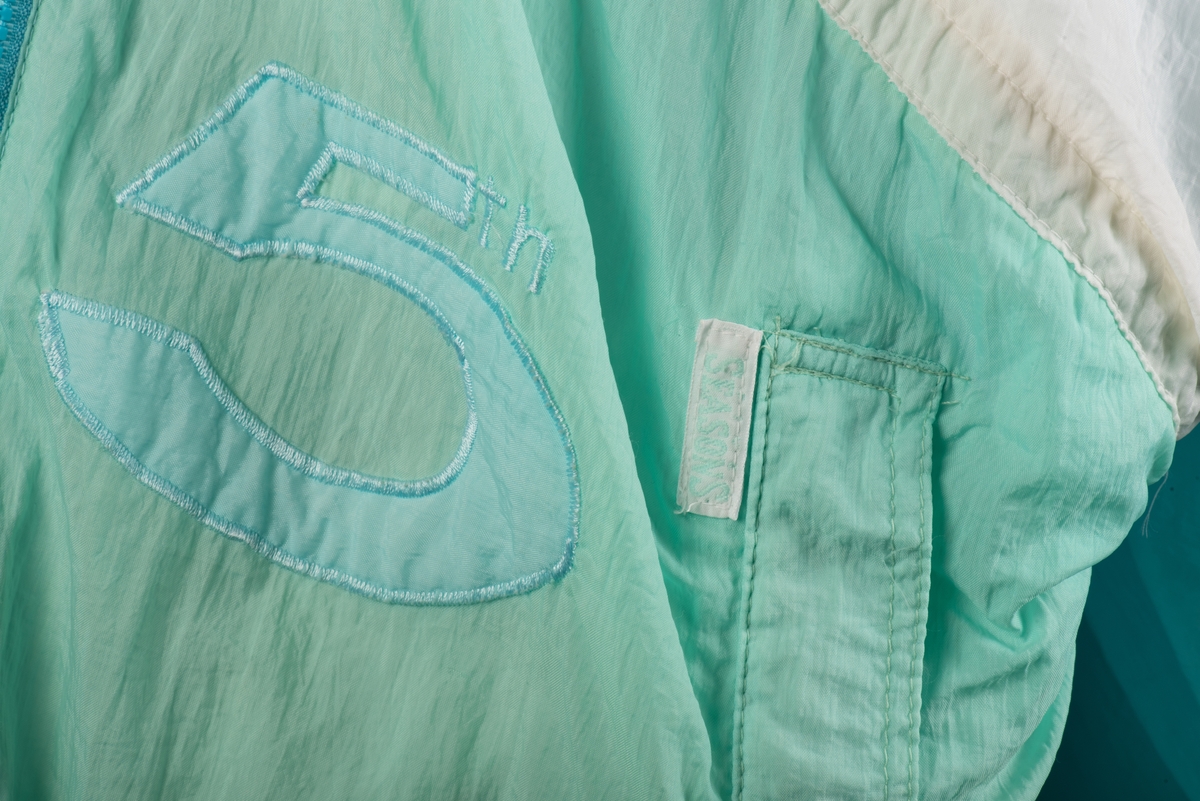 Skidjacka i pingvinmodell tillverkad av syntetmaterial, nylon/polyester.
Jackan i turkost, rosa, mintgrönt och vitt är sydd i så kallade pingvinmodell. Mönstret består av oregelbundna fält med de olika färgerna. På framsidan finns ditsydda applikationer med siffrorna 1 till 5 i olika färger. Den ljusblå dragkedjan är placerad något till vänster och diagonalt på framsidan. Den upprättstående kragen är försedd med två tryckknappar, och innanför finns ytterligare en krage. Det finns två fickor, försedda med dragkedjor. Jackan har insydda resårer nedtill samt i ärmsluten. Nedtill finns det även en dragsko med snodd.