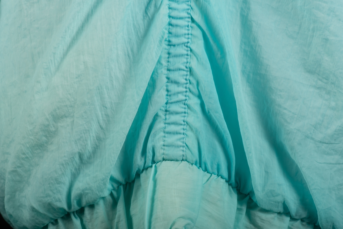 Skidjacka i pingvinmodell tillverkad av syntetmaterial, nylon/polyester.
Jackan i turkost, rosa, mintgrönt och vitt är sydd i så kallade pingvinmodell. Mönstret består av oregelbundna fält med de olika färgerna. På framsidan finns ditsydda applikationer med siffrorna 1 till 5 i olika färger. Den ljusblå dragkedjan är placerad något till vänster och diagonalt på framsidan. Den upprättstående kragen är försedd med två tryckknappar, och innanför finns ytterligare en krage. Det finns två fickor, försedda med dragkedjor. Jackan har insydda resårer nedtill samt i ärmsluten. Nedtill finns det även en dragsko med snodd.