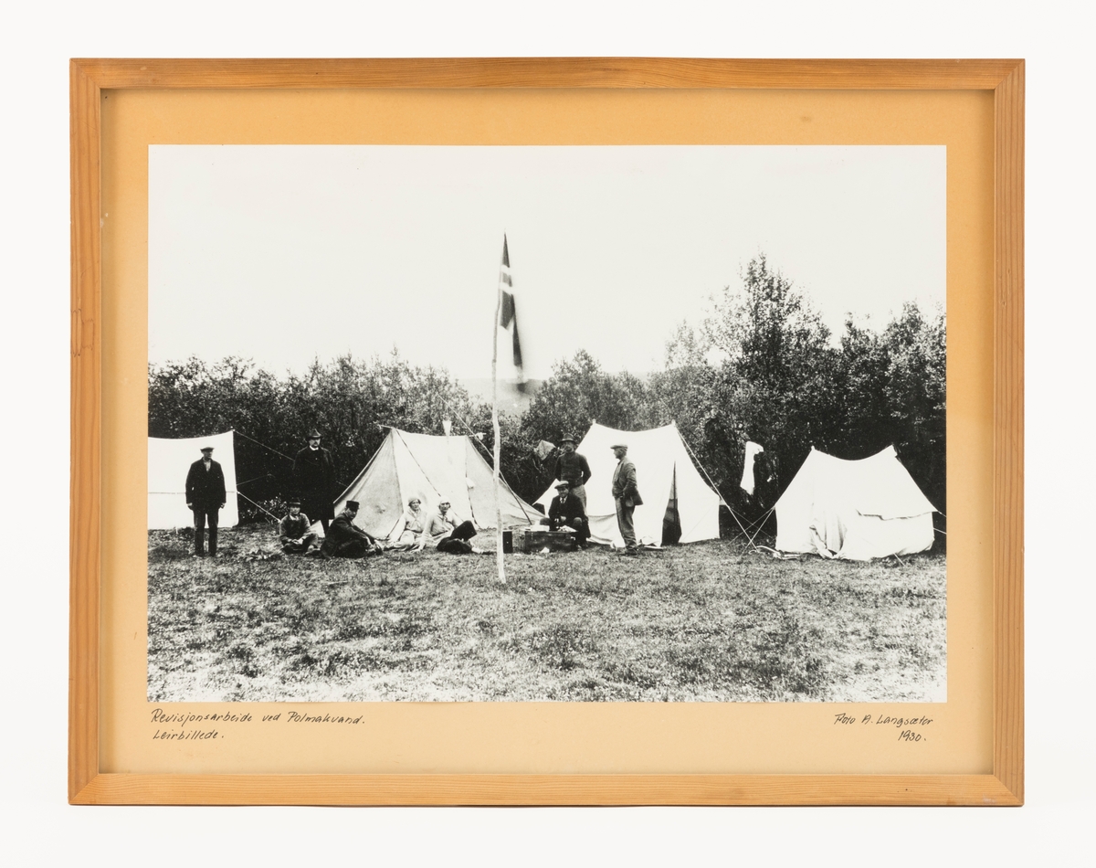 Fotografi fra teltleir som ble etablert ved Polmakvannet/Buolbmátjávri under revisjonsarbeid i 1930. Dette arbeidet ble utført som et ledd i Det norske skogforsøksvesens program for «Måling og beregning av skogforråd og tilvekst». Ambisjonen var å utarbeide kubikk- og tilveksttabeller for skogbruket i ulike deler av landet. Dette innebar mye feltarbeid sommerstid, ofte med enkle innkvarteringsforhold for personalet. Her var teltene plassert på ei grasslette omgitt av bjørkekratt. Sentralt på sletta var det reist en staur med et norsk flagg. Ved slettas ytterkanter sto det fire telt. Mellom teltene sto og satt det det ni personer, sju menn og to kvinner.