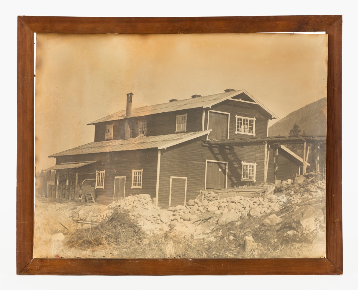 Svart-hvitt-fotografi av Hanestad tjærefabrikk i Øvre Rendalen. Denne virksomheten ble drevet av skogeieren Andreas Mohr Øvergaard på garden Østre Hanestad i Rendalen og kollegaen Otto Aakrann, som bodde på Gårder i Elverum i perioden 1911-1920. Produksjonsanlegget lå på førstnevntes eiendom. Det besto av en bygning som later til å ha vært reist i bordkledd bindingsverk. Midtpartiet hadde to etasjer og bølgeblikktekket saltak. I dette taket var det tre murte piper - antakelig fra tjæreovnene (på baksida i forhold til hvor dette bildet er tatt). Langs begge langveggene var det enetasjes utbygg med pulttak. Fra det utbygget som lå nærmest fotografen da dette bildet ble tatt gikk det en luftekanal oppover langs veggen mot den toetasjes midtfløyen. Sannsynligvis var det i utbygget på denne sida motorene som drev maskinene i fabrikken sto. Til andre etasje i midtfløyen gikk det ei kjørebru av tre. Antakelig var det der råstoffet til produksjonen ble hentet inn.  Det lå en del stein framfor bygningen. Dette var muligens stein som var ryddet til side i forbindelse med bygginga av fabrikken.