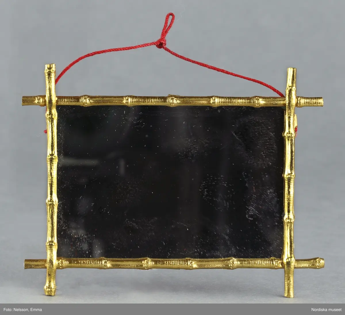 Inventering Sesam 1996-1999:
H 8,3 cm
B 10,7 cm
Dockskåpsspegel, väggspegel, fyrkantig ram av förgylld metall imiterande bambu. Spegelglas fodrat med rött papper på baksidan. Spegelglaset fäst med små metalltungor. Upphängningsanordning  av rött garn.
Tillhör dockskåp inv 164.570. Brukad av givarinnan född 1886.
Leif Wallin nov 1997