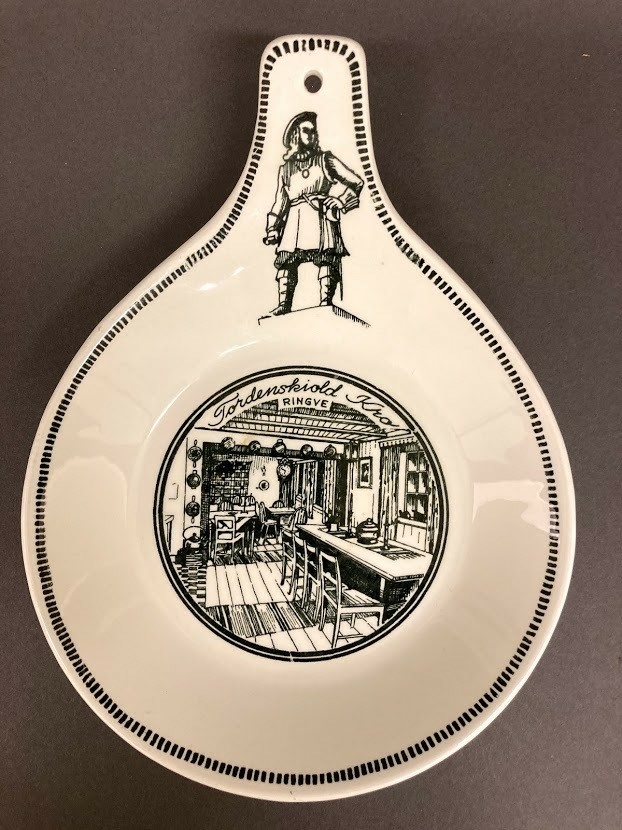 Avlang skål med håndtak, hull til oppheng. Ment for å henge på vegg. Dekorert på håndtak med helfigur av Tordenskiold. I skålens bunn dekorert med bilde av interiøret i "Tordnskiold-kro",