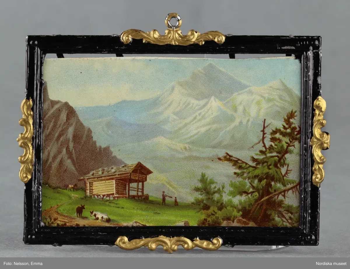 Inventering Sesam 1996-1999:
L 8,7 cm
B 6,3 cm
3 st dockskåpstavlor, flerfärgstryck på papp, tunn ram av tenn, svartmålad med guldmålade ornament.
a) Motiv; alplandskap med litet hus.
b) Motiv; by i alplandskap med kyrka, text i nedre högra hörnet "St Maurice".
c) Motiv; borg på bergssluttning, text i nedre högra hörnet "Rheinstein".
Tillhör dockskåp inv 164.570. Brukad av givarinnan född 1886.
Leif Wallin nov 1997