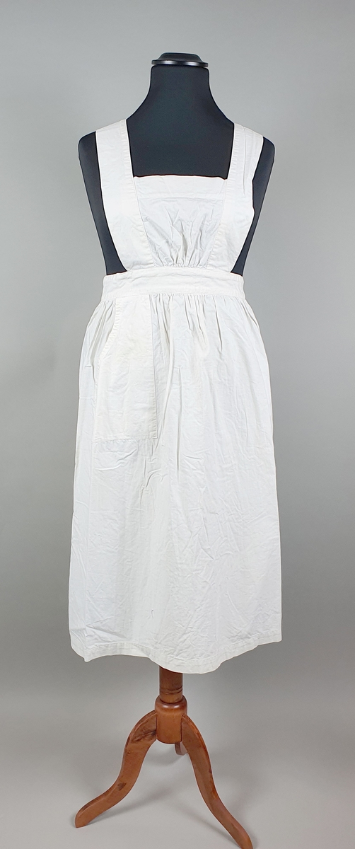 Hvitt forkle av bomull for sykepleier. Forkleet har smekke med bånd som festes diagonalt over ryggen med knapp i linningne. En påsydd lomme midt foran.