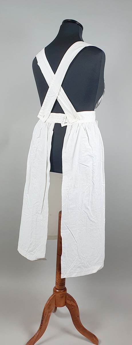 Hvitt forkle av bomull for sykepleier. Forkleet har smekke med bånd som festes diagonalt over ryggen med knapp i linningne. En påsydd lomme midt foran.