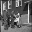 Hemvärnsmän och lottakår vid ett bord med fika utanför en okänd större byggnad med locklistpanel. En kvinna häller upp kaffe till en hemvärnsman. En ung man med pipa i munnen vänders sig mot fotografen.