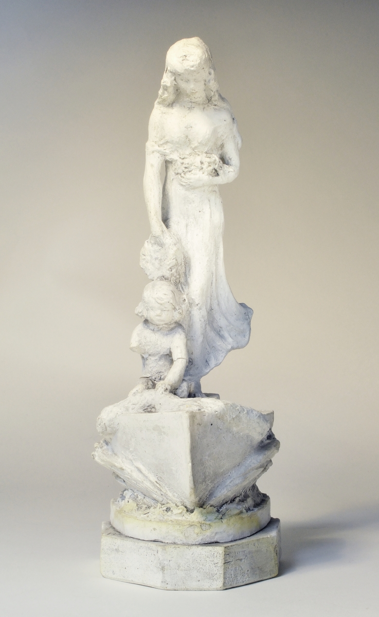 Skulpturskiss av Ida Matton, signerad Paris 1920, till gravmonument för redarfamiljen Brodin i Gävle. Kvinna med blommor i händerna med ett barn med blomgirlang sittande framför fötterna. Båda placerade på en trekantig sockel som liknar stäven på en båt.
