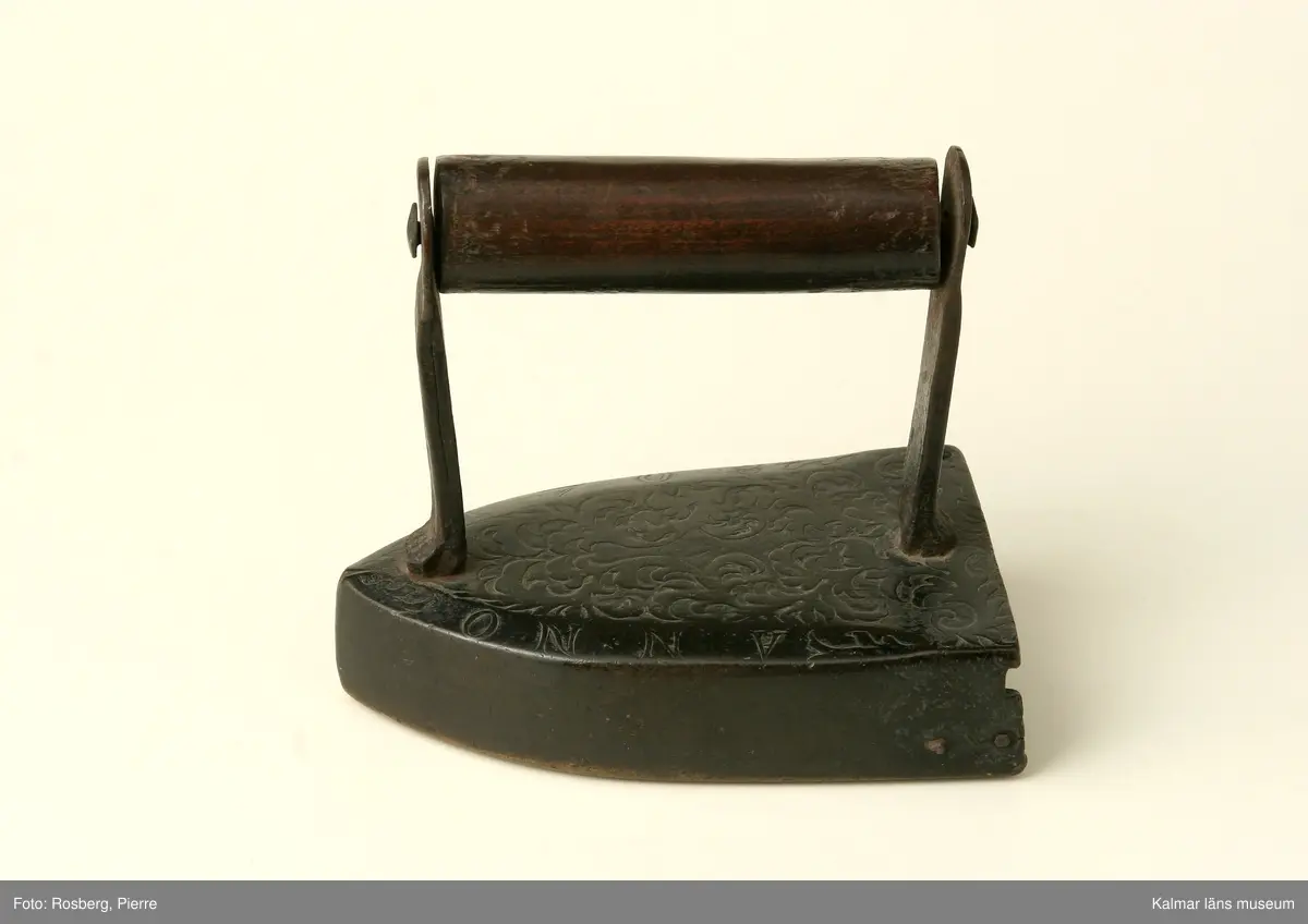 KLM 5925. Strykjärn. Av malm. Järnet framtill spetsigt. Handtag av trä. Lodet och luckan saknas. Övre ytan prydd med gjuten reliefornering i form av rankor samt ANNO 1708 .