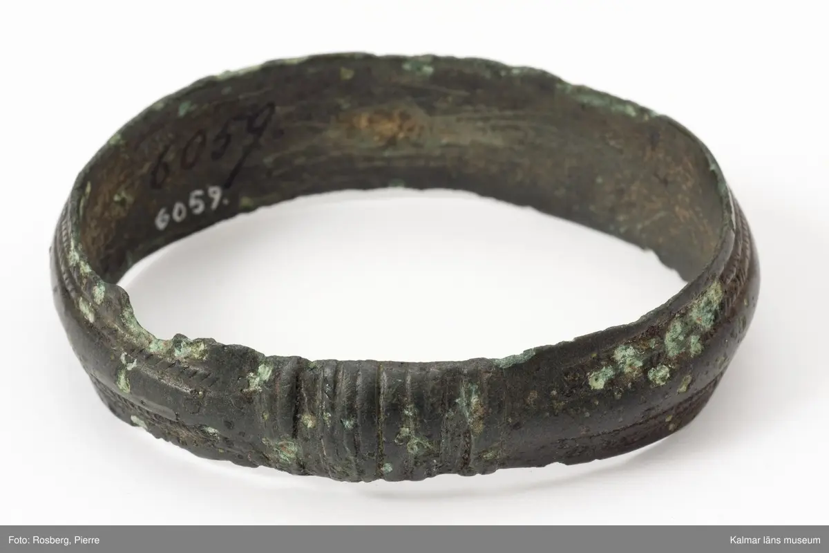 KLM 6059. Armring av brons. Nästan jämnbred och sluten. Smala åsar längs med ringen, två på var sida, är snett streckade tätt intill varandra med 12-13 tvärsträck växelvis, imiterande en omvirning.