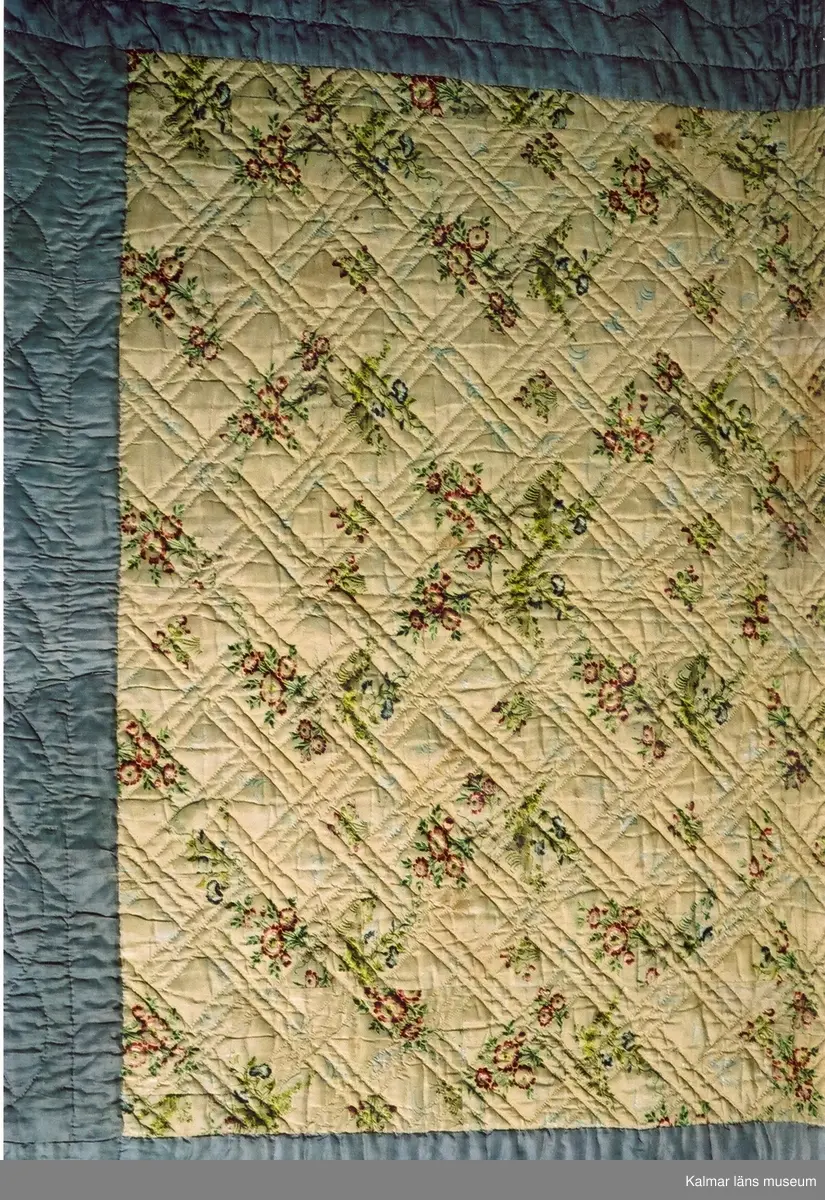 KLM 9174. Täcke, troligen brudtäcke. Spegel av ljusgult, mönstervävt siden med orientaliskt inspirerat mönster av blommor och blommande träd i rosa, rött, blått, lila grönt och beige. Bård av grålila siden, senare påsydd. Sidenet har många skarvar. Foder av oblekt linne. Vaddfyllning. Sticksöm utförd i lingarn i ett diagonalt rut- eller flätmönster. Längs kanterna på det ursprungliga täcket en smal bård av löv samt vid ena kortsidan årtalet 1797 och ett flertal initialer: EH, DH, PHDHW, SH, FH.  Den senare påsydda grålila bården har sticksöm utförd i vit bomullstråd i mönster av överlappande cirklar och med fyrbladiga blommor i hörnen. På bården påskrift med märkbläck.1893. Lagat. M.B. Fälla, (eller M.R. Tälla).