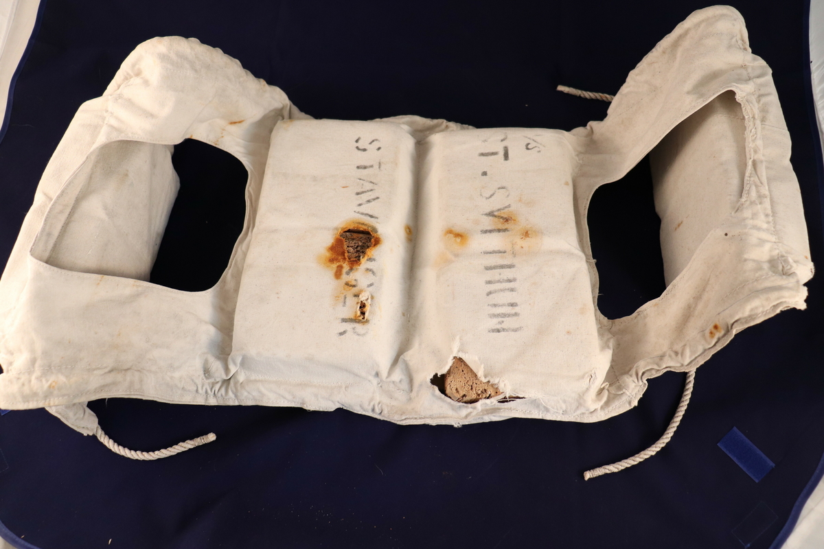 Standard redningsvest til å knyte rundt livet. Vesten er laget av seilduk og flyteposene er fylt med kork. Har noen hull på posene med noe rustfarge rundt, teksten som er trykt på er nesten helt borte.