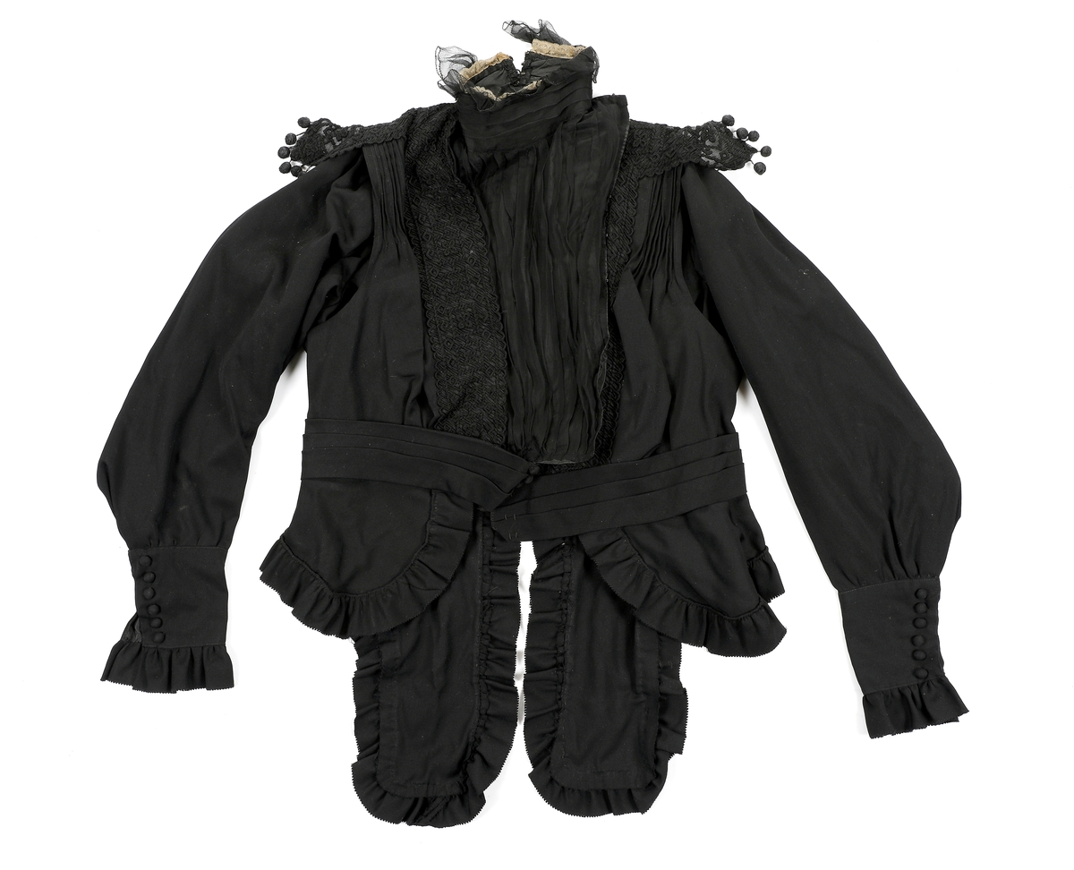 Helforet med svart mugg, 10 fine legg på forstykke som er løst. Bryststykke og halslinning har foldelagt chiffon. Øverst på halslinning rynkekappe av chiffon og smal hvit bobinetteblonde. Brodert chiffonlengde fra halsen og ut over skuldra. Svart blonde ut på skuldrene og på ned i en spiss V foran. Belte i livet. Kappe på hver side under beltet med volang. Kappene fortsetter ned i to snipper bak. Mansjettene på hver erme er høye og lukkes med trykknapper med pynteknapper over.
 