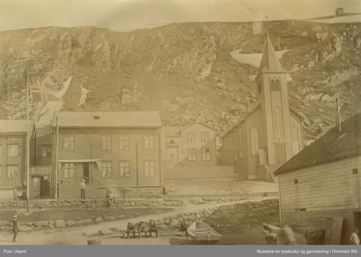 Den katolske kirken i Hammerfest i 1899. Til venstre for den står det trehus med skifertak. Foran husene står det en flagmast med et vaiende  norgesflagg. Nede i midten av bildet står en liten båt og ved siden av sitter tre men på en benk. Til venstre for dem er det en uniformert person. Arbeidsfolk er å se ved pakkhuset. Oppe på veien står det en kvinne og to barn. En man sitter med et barn i fanget på muren. Det virker som om innbyggerne i Hammerfest nyter godværet.  I fjellsida til Salen ligger det fortsatt litt sne. Oppe på Salenfjellet ser man varden.
