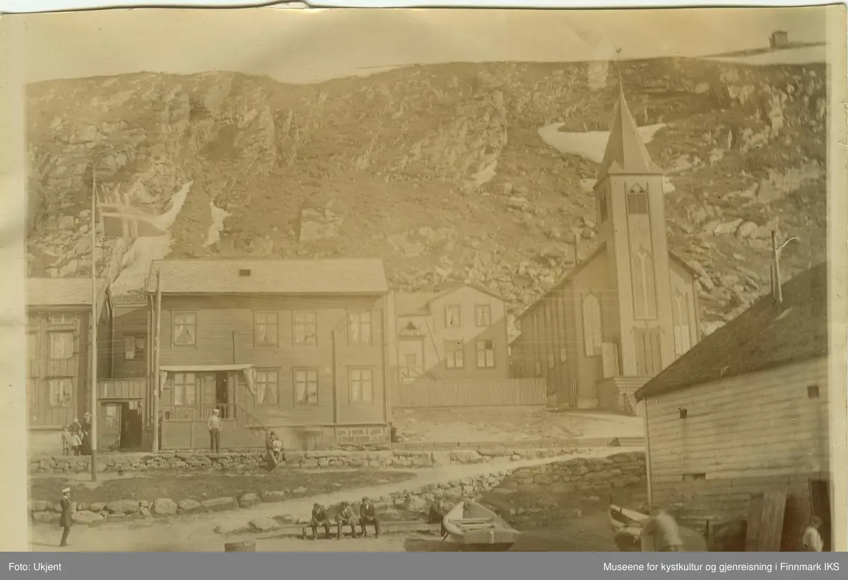 Den katolske kirken i Hammerfest i 1899. Til venstre for den står det trehus med skifertak. Foran husene står det en flagmast med et vaiende  norgesflagg. Nede i midten av bildet står en liten båt og ved siden av sitter tre men på en benk. Til venstre for dem er det en uniformert person. Arbeidsfolk er å se ved pakkhuset. Oppe på veien står det en kvinne og to barn. En man sitter med et barn i fanget på muren. Det virker som om innbyggerne i Hammerfest nyter godværet.  I fjellsida til Salen ligger det fortsatt litt sne. Oppe på Salenfjellet ser man varden.