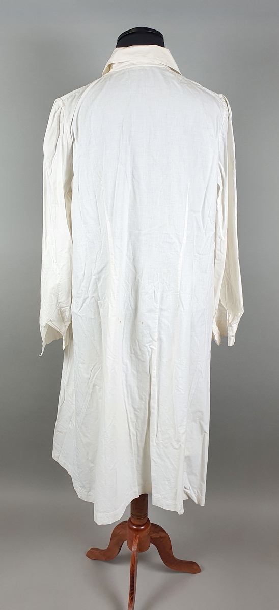 Hvit sykepleiefrakk for kvinne. Skjortekrage, lange ermer med mansjett og knapper av kartong midt foran, og en brystlomme på venstre forstykke og to påsydde lommer på hvert forstykke. Frakken er noe flekkete.