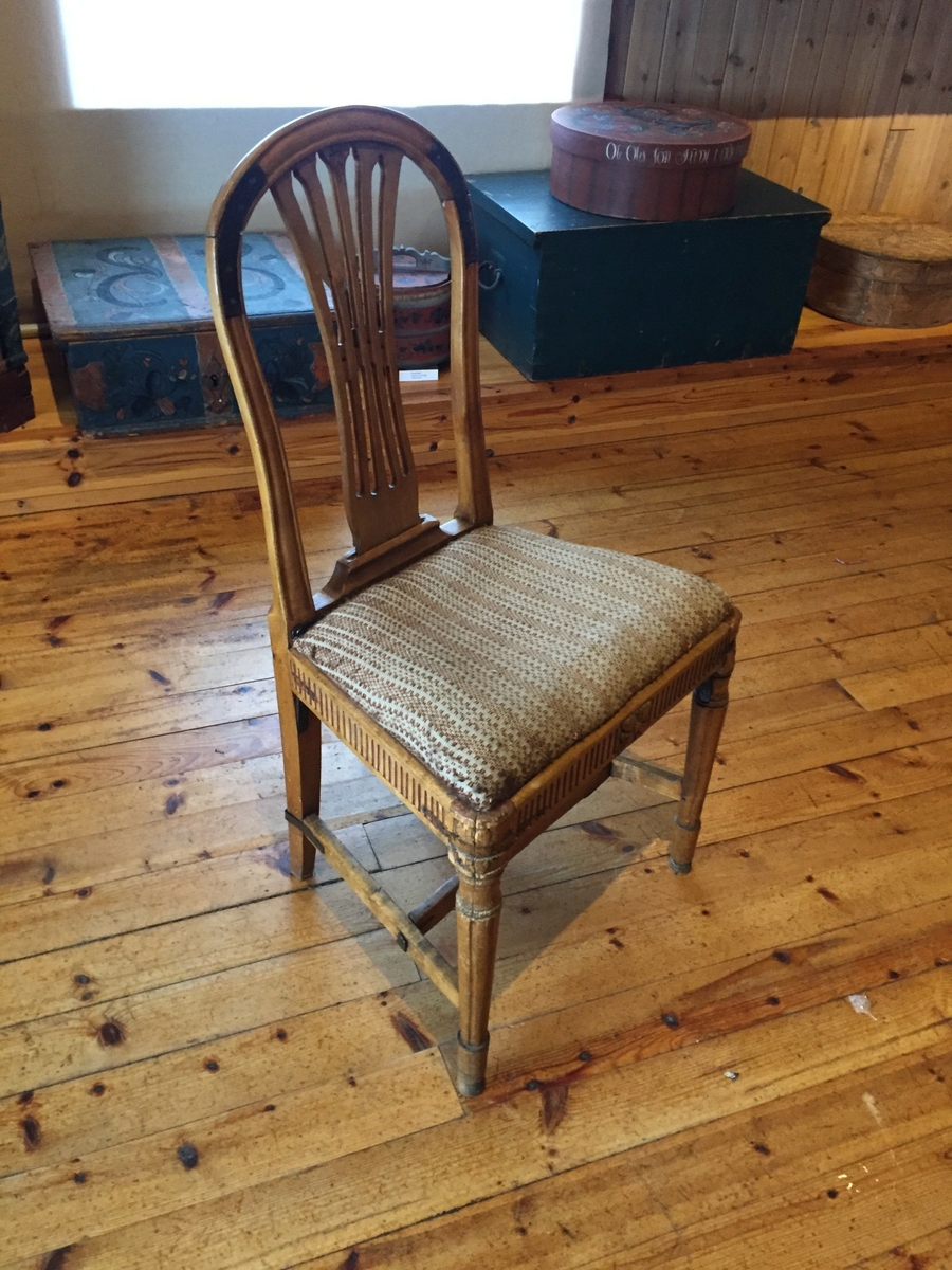 Stol med boga rygg. Spiler med utskjeringar i ryffen. Utskjeringar kring setet. Beina framme er dreia. Pute med heimevove trekk. Puta er laus. Det er eldre stoff (ullstoff) under det trekket som er på nå.
