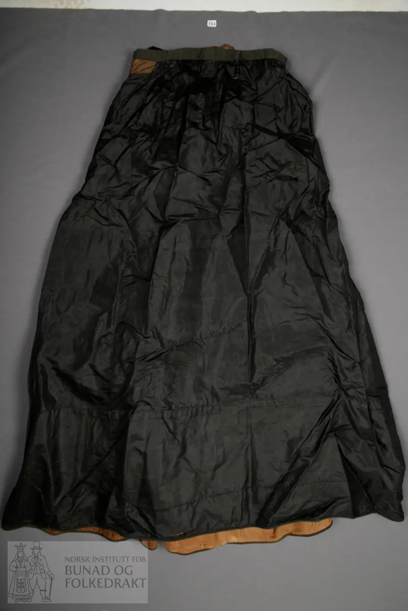 Brudeskjørt i svart silketaft, trolig brukt sammen med understakk NBF2021-0840.
Mørk grønt kypertvevd bånd påsydd for hånd rundt nedkant. 
Bomullsstoff , lerret til skoning.
Piperynket bak.
Splitt litt mot venstre side bak.