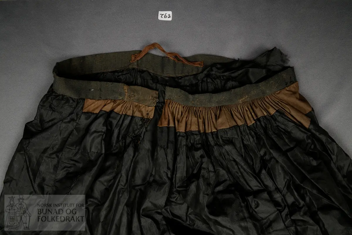 Brudeskjørt i svart silketaft, trolig brukt sammen med understakk NBF2021-0840.
Mørk grønt kypertvevd bånd påsydd for hånd rundt nedkant. 
Bomullsstoff , lerret til skoning.
Piperynket bak.
Splitt litt mot venstre side bak.