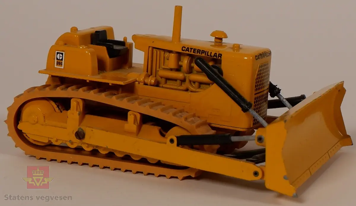 Caterpillar D9G. Leketøy, miniatyrbulldozer av metall og gummi. Gul med enkelte svarte detaljer. Dozeren går på belter og har skjær foran. Merking fra produsenten under leketøyet samt Caterpillar merking utenpå.