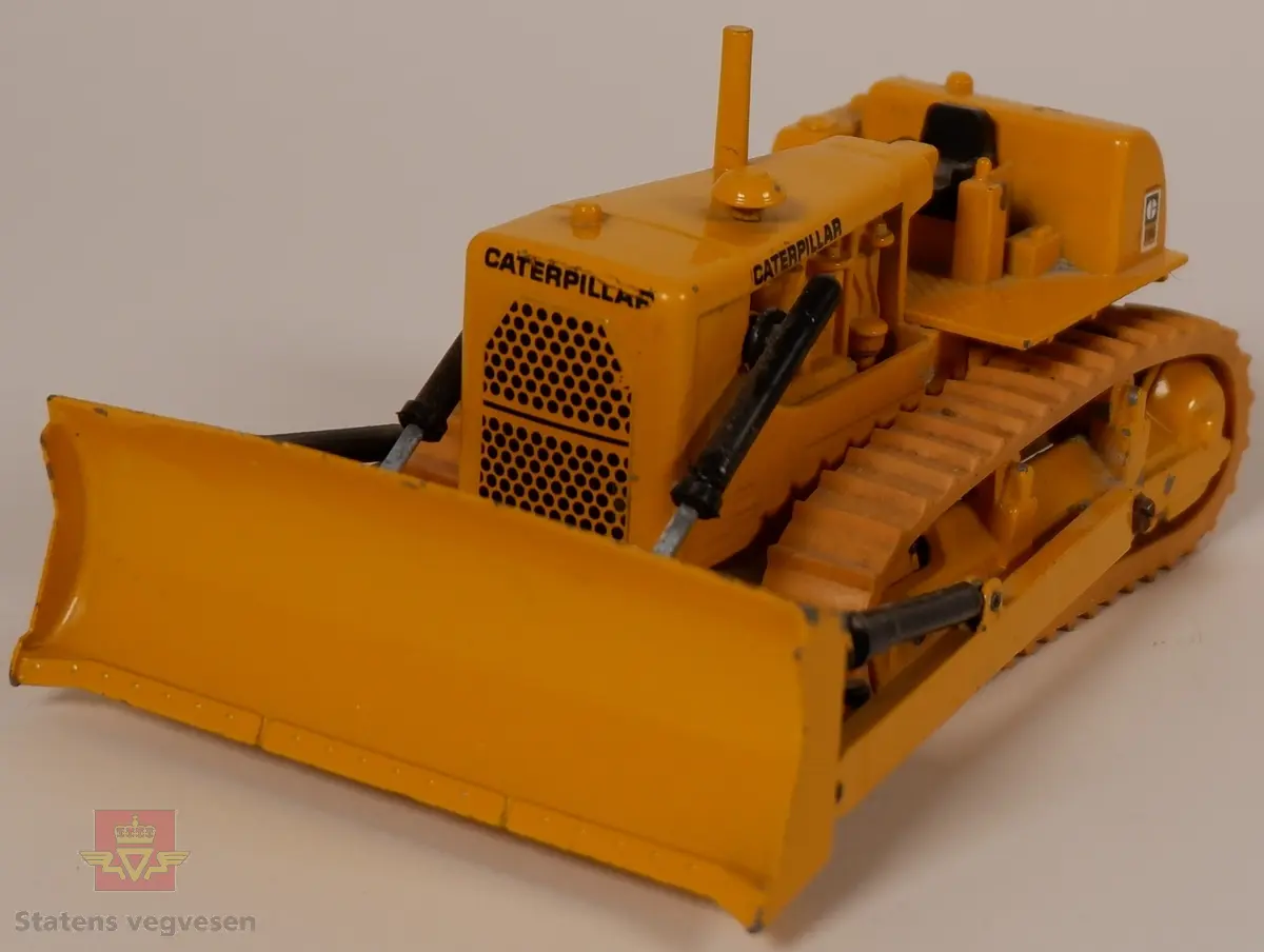 Caterpillar D9G. Leketøy, miniatyrbulldozer av metall og gummi. Gul med enkelte svarte detaljer. Dozeren går på belter og har skjær foran. Merking fra produsenten under leketøyet samt Caterpillar merking utenpå.