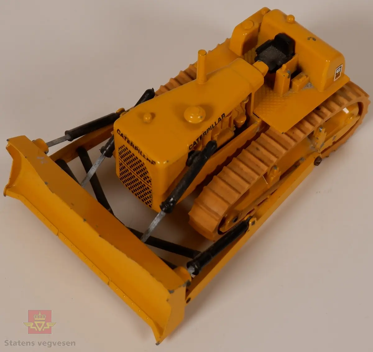 Caterpillar D9G. Leketøy, miniatyrbulldozer av metall og gummi. Gul med enkelte svarte detaljer. Dozeren går på belter og har skjær foran. Merking fra produsenten under leketøyet samt Caterpillar merking utenpå.
