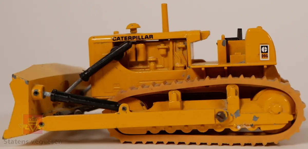 Caterpillar D9G. Leketøy, miniatyrbulldozer av metall og gummi. Gul med enkelte svarte detaljer. Dozeren går på belter og har skjær foran. Merking fra produsenten under leketøyet samt Caterpillar merking utenpå.