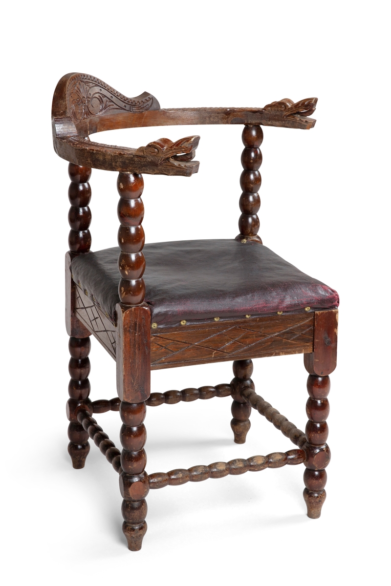 Trestol med diagonalt plassert, firkantet sete med mørkerød voksduk og blomsterformede nagler. Med kuleformer langs bein, sprosser, samt vertikal ryggdel. Øvre del av ryggen er halvsirkelformet og går over til armlenene som har et utskjært dragehode i hver ende.