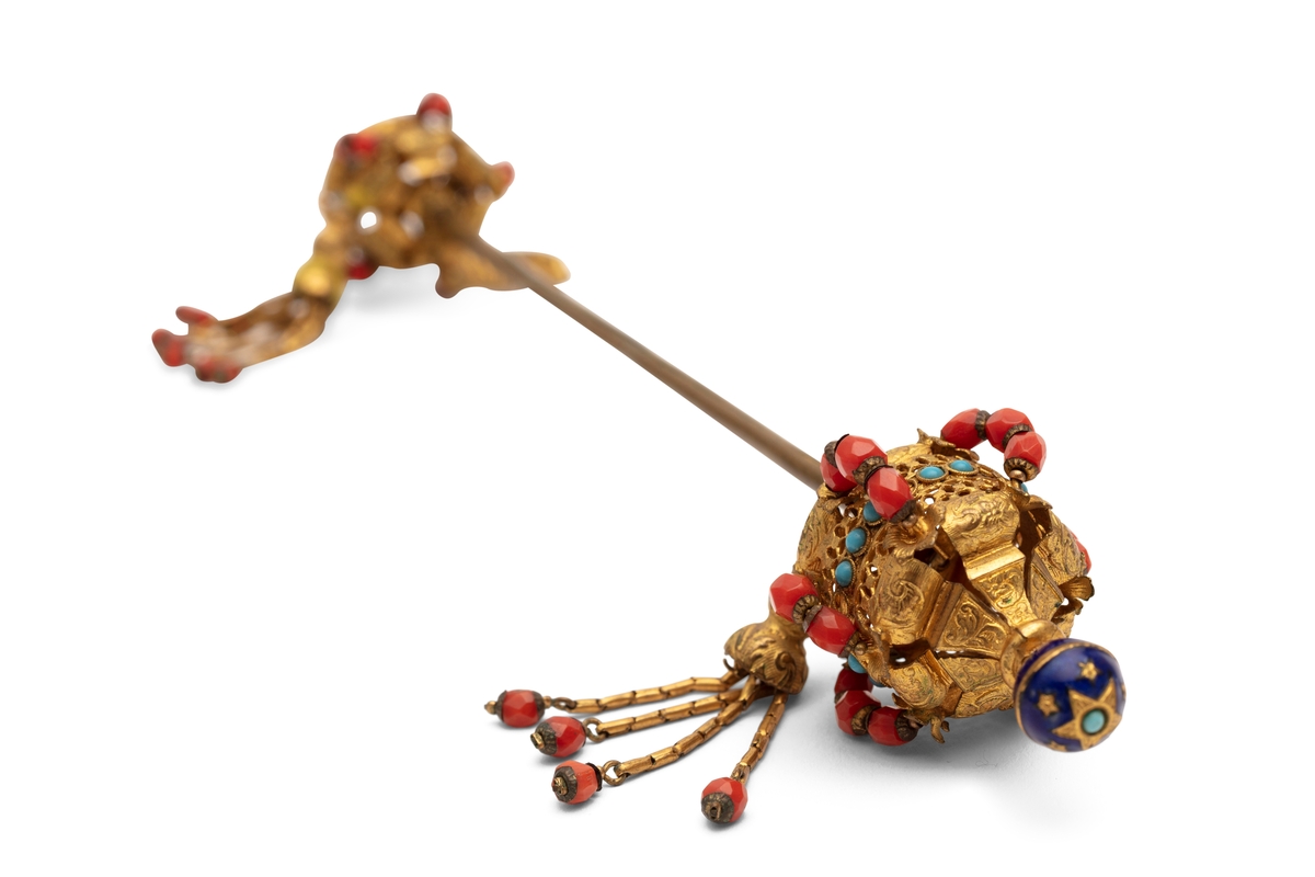 Hattenål i metall, delvis forgylt. Endestykkene er dekorert med røde og blå glassperler og dusker.