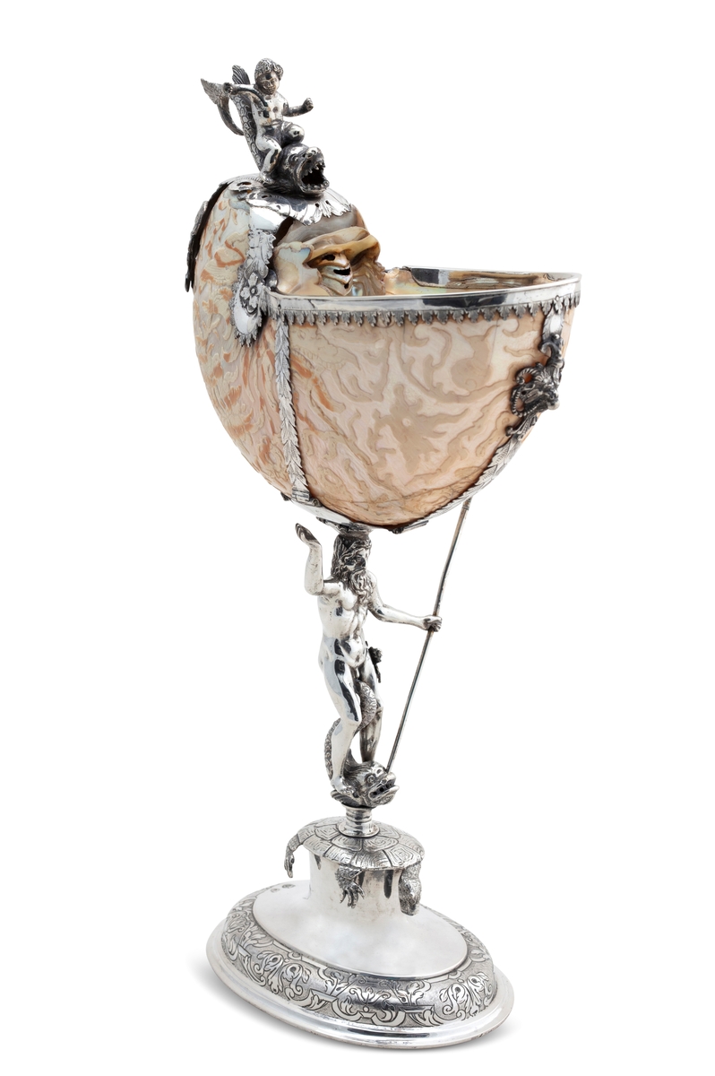 Skall av blekksprutarten Nautilus. Skåret dekorasjon, fot og innfatning sølv, støpt og gravert dekorasjon. Stpl: MF/H//1720 Morten Finchenhagen, Christiania. Skallet er brunflammet i overflaten. Nautilusblekkspruten lever i sydlige hav omkring Filippinene.