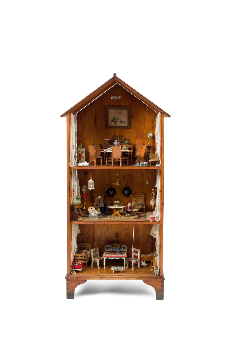 Dukkehus i tre. Huset har 3 etasjer og et spisstak. Hver etasje har 2 vinduer med gardiner. Overflaten er beiset og lakkert.