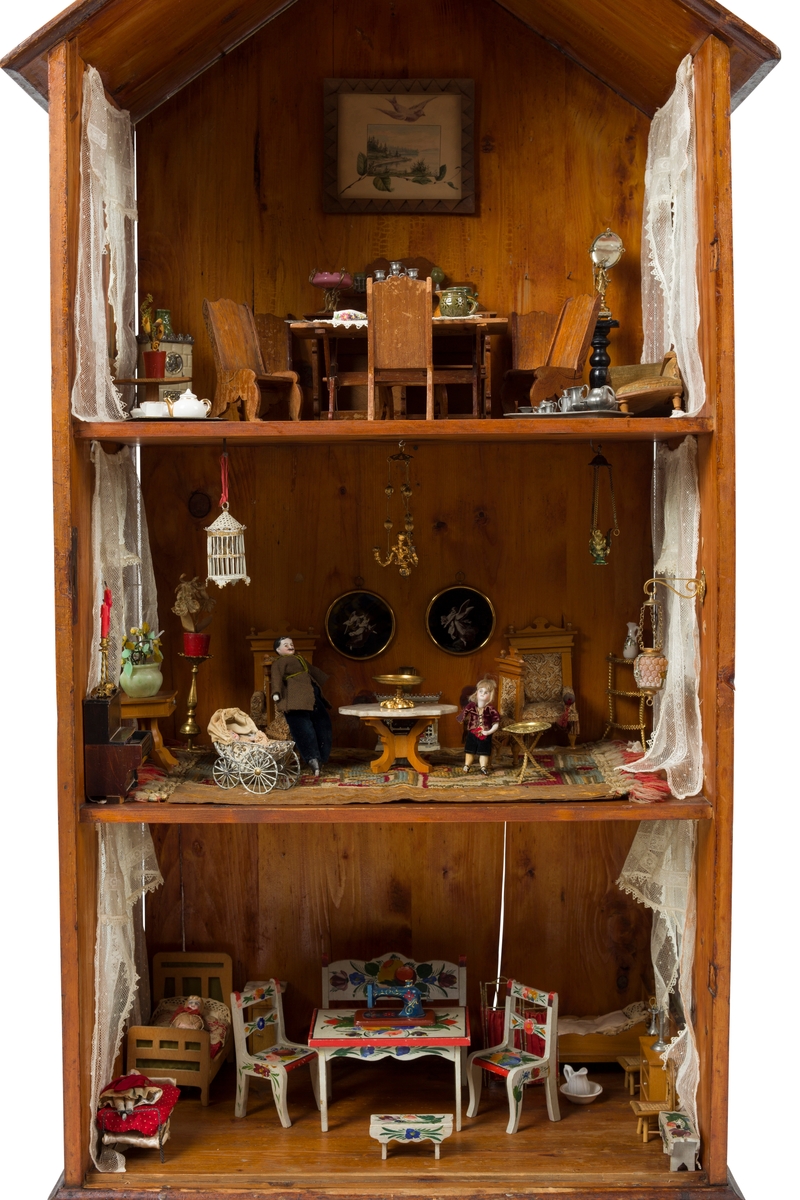 Dukkehus i tre. Huset har 3 etasjer og et spisstak. Hver etasje har 2 vinduer med gardiner. Overflaten er beiset og lakkert.