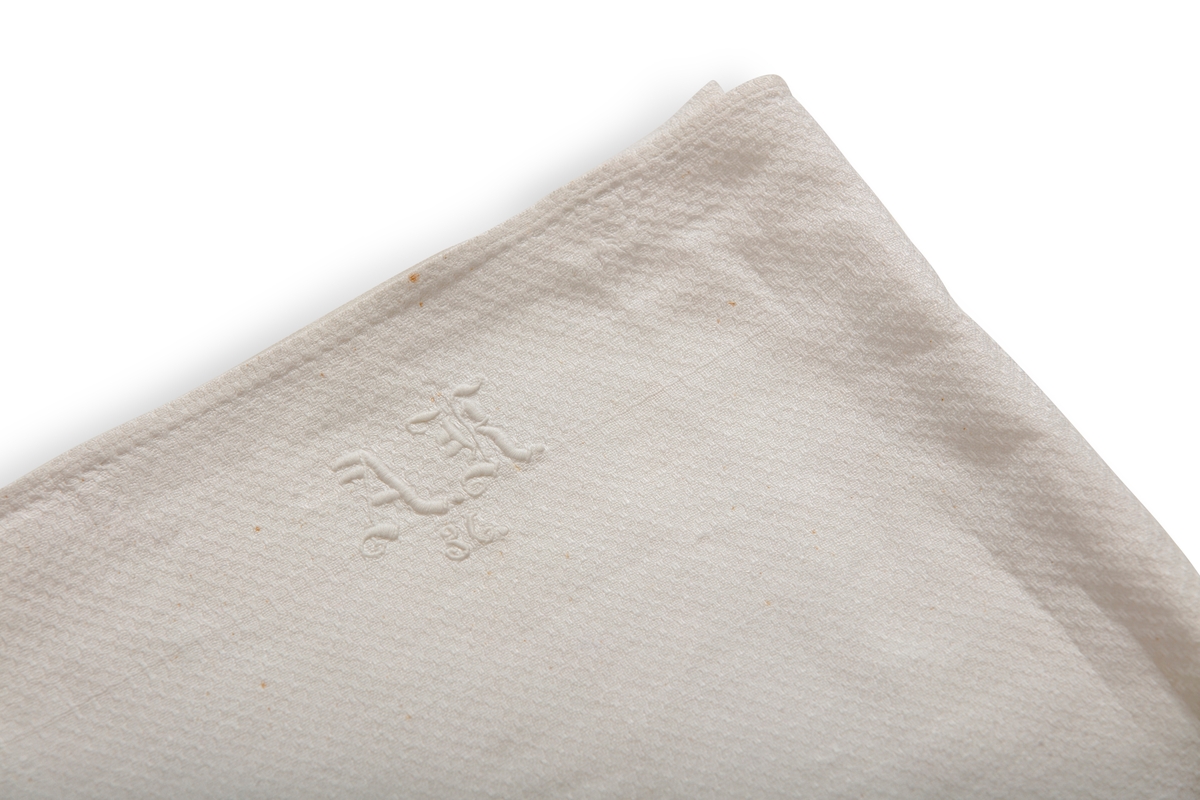Håndkle av hvit lin med mønstereffekt i vevningen. Maskinsydd fold i hver ende. Hvitt håndbrodert monogram i plattsøm. Påsydd hank av bomullsbånd.