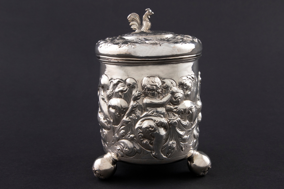 Drikkekanne i sølv med sylinderformet korpus, hank og hengslet lokk. Kannen står på tre kuleføtter dekorert med hver sin blomst. Korpus er dekorert med drevet blomsterdekor blant tre kjeruber. Lokket har en krans med drevet blomster- og akantusranke med fugler. I sentrum er en sirkel med initialer inni en laurbærkrans. Lokkåpner er formet som en hane. Hanken er dekorert med akantusranke som ender i en blomst.