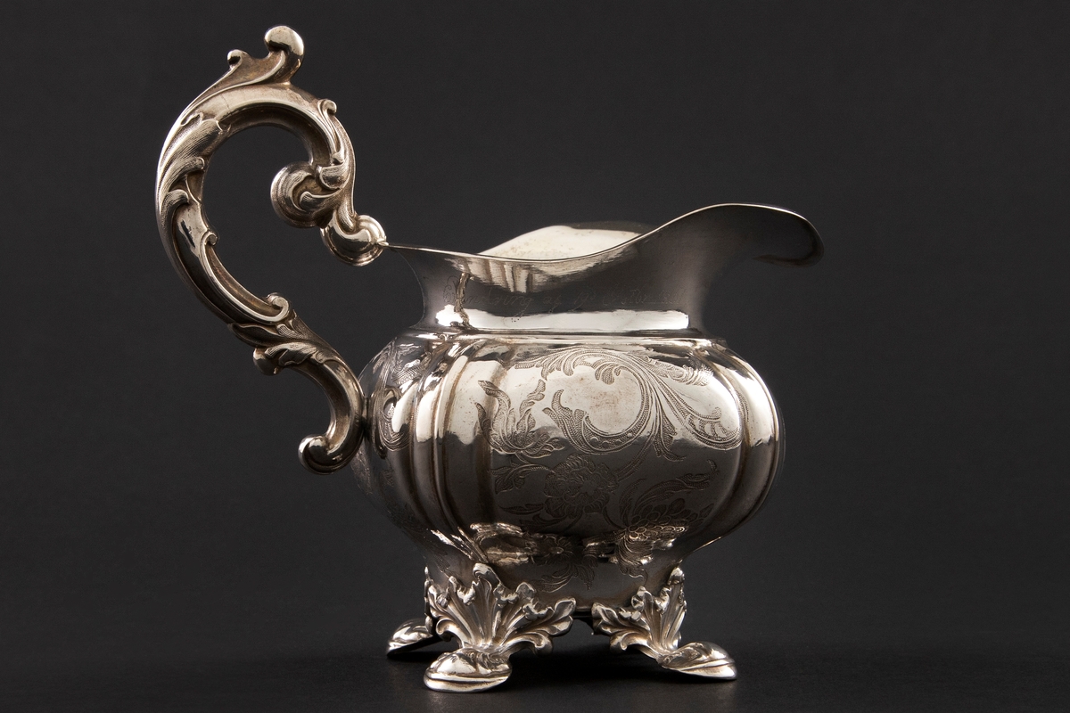 Fløtemugge i nyrokokko i presset sølvblikk, dekorert med presset gravyr i form av blomster- og bladornamenter. Muggens hviler på fire påloddede bein, tilnærmet kvadratisk utbulet korpus, samt bred helletut. S-formet hank i nyrokokko-ornamentikk.