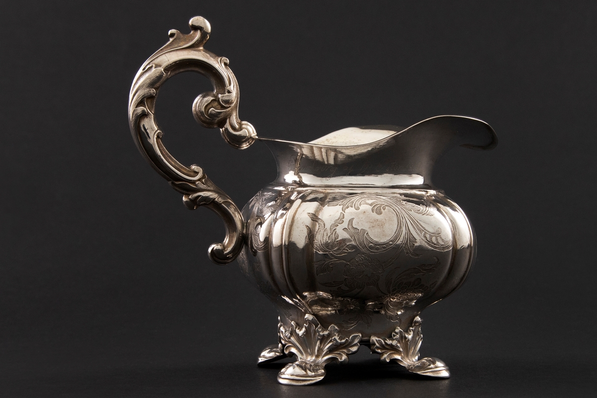 Fløtemugge i nyrokokko i presset sølvblikk, dekorert med presset gravyr i form av blomster- og bladornamenter. Muggens hviler på fire påloddede bein, tilnærmet kvadratisk utbulet korpus, samt bred helletut. S-formet hank i nyrokokko-ornamentikk.