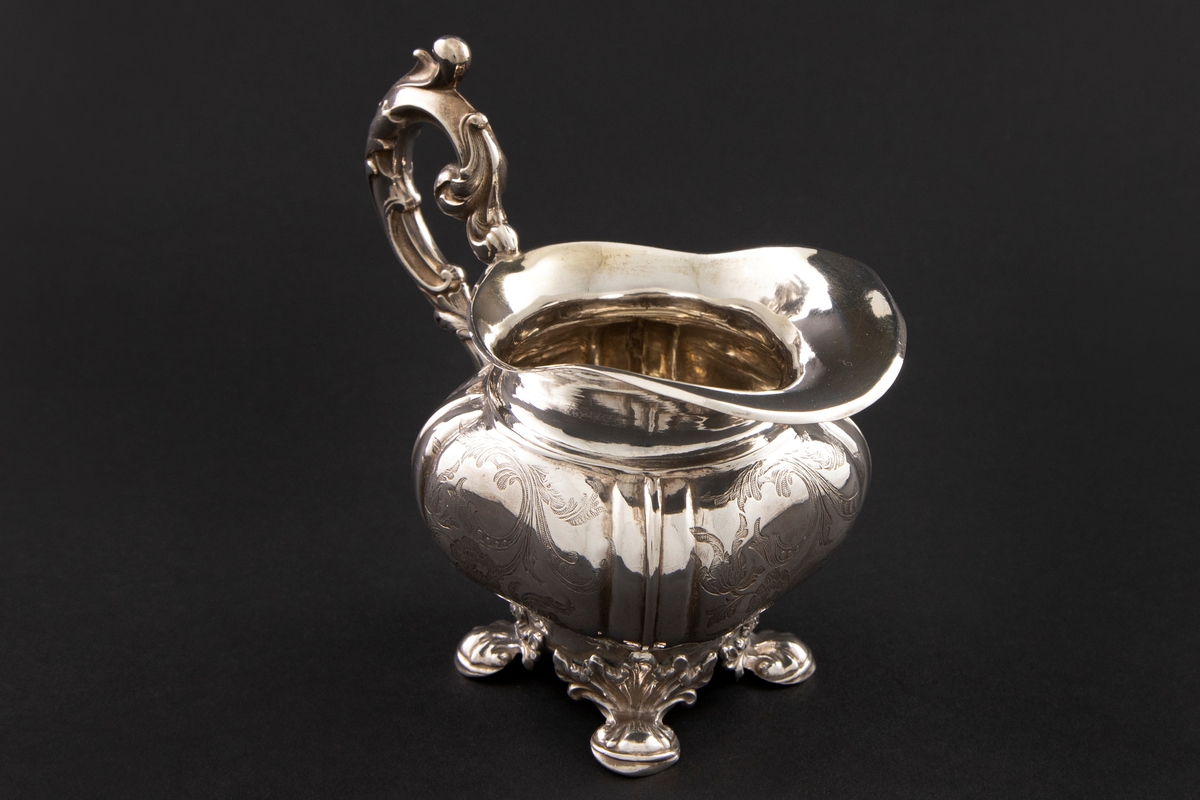 Fløtemugge i nyrokokko i presset sølvblikk, dekorert med presset gravyr i form av blomster- og bladornamenter. Muggens hviler på fire påloddede bein, tilnærmet kvadratisk utbulet korpus, samt bred helletut. S-formet hank i nyrokokko-ornamentikk.