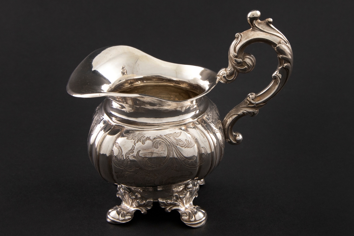 Fløtemugge i nyrokokko i presset sølvblikk, dekorert med presset gravyr i form av blomster- og bladornamenter. Muggens hviler på fire påloddede bein, tilnærmet kvadratisk utbulet korpus, samt bred helletut. S-formet hank i nyrokokko-ornamentikk.