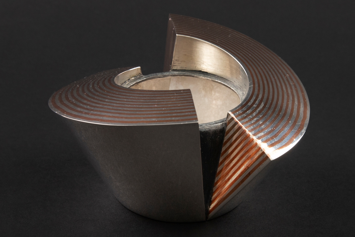 Eske i sølv, hvis korpus preges av et stramt geometrisk formspråk bestående av to halvsirkelformede deler, som omslutter et sylinderformet midtparti. Overflaten er delvis dekorert med striper i kobber som er loddet sammen med sølvet. Mellomrommene mellom delene er oksidert.