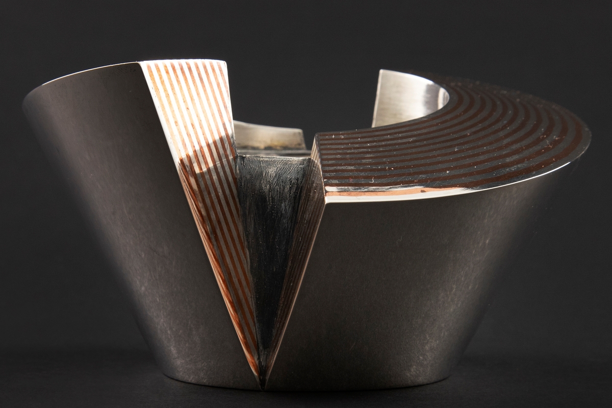 Eske i sølv, hvis korpus preges av et stramt geometrisk formspråk bestående av to halvsirkelformede deler, som omslutter et sylinderformet midtparti. Overflaten er delvis dekorert med striper i kobber som er loddet sammen med sølvet. Mellomrommene mellom delene er oksidert.