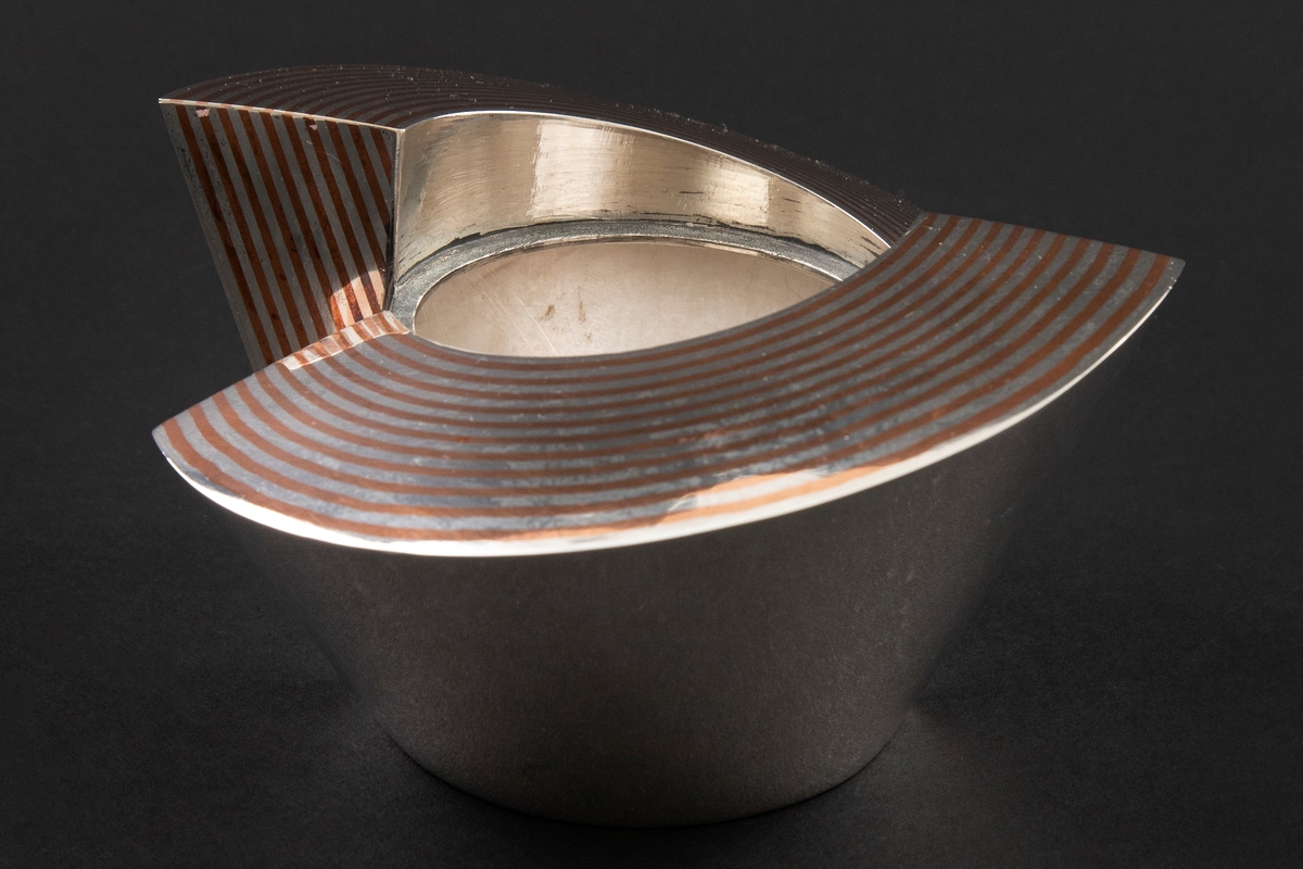 Eske i sølv, hvis korpus preges av et stramt geometrisk formspråk bestående av to halvsirkelformede deler, som omslutter et sylinderformet midtparti. Overflaten er delvis dekorert med striper i kobber som er loddet sammen med sølvet. Mellomrommene mellom delene er oksidert.