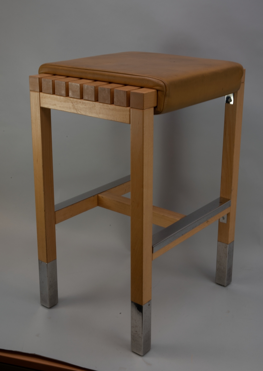 Hög sittpall i stil med en barstol, av ljust trä med fyrkantig ribbad sits med fast dyna klädd med ljusbrunt konstläder. Pallen har kromad förstärkning nertill på alla benen, samt på tvärslåarna nertill där man kan placera fötterna.
