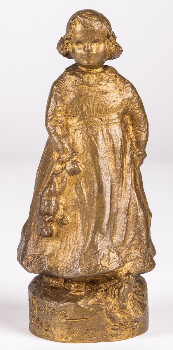 Skulptur eller statyett av brons, "En solstråle" av Ida Matton. Signerad Ida Matton, Paris. Liten flicka med lockigt hår, fotsid klänning med rosett bak. Håller en nallebjörn i höger hand. En docka ligger framför vänstra foten.