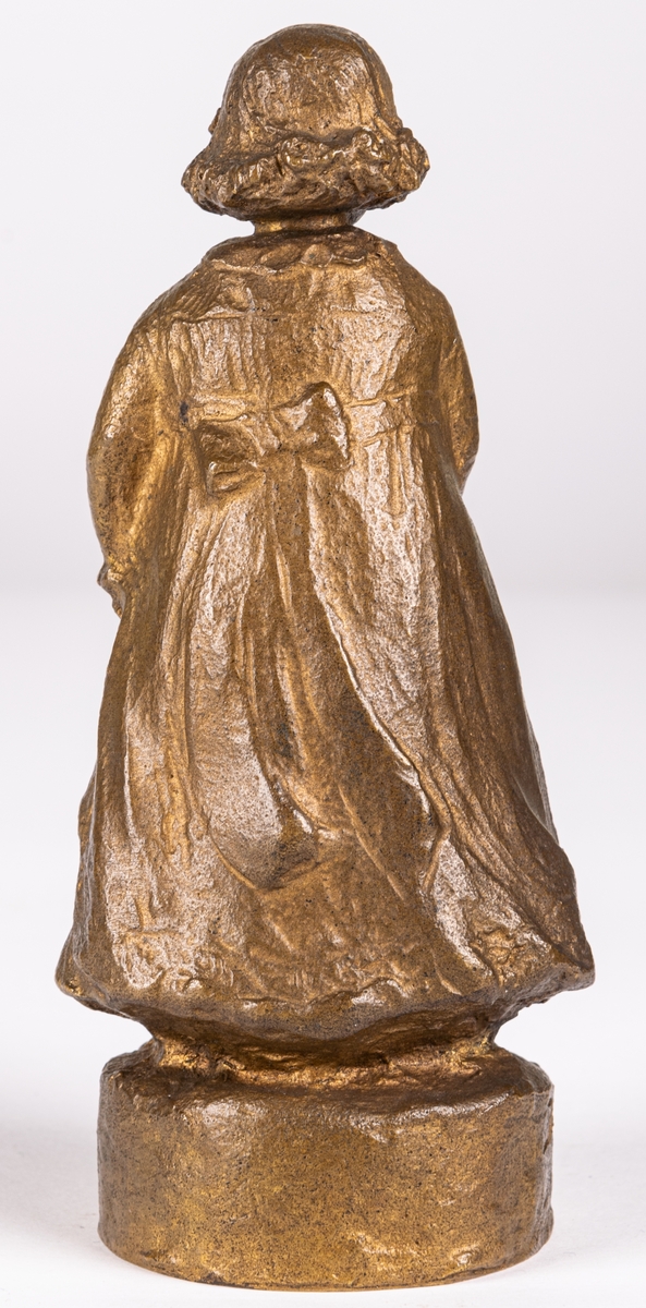 Skulptur eller statyett av brons, "En solstråle" av Ida Matton. Signerad Ida Matton, Paris. Liten flicka med lockigt hår, fotsid klänning med rosett bak. Håller en nallebjörn i höger hand. En docka ligger framför vänstra foten.