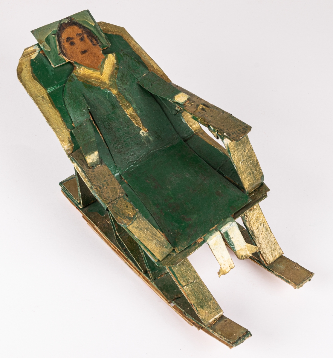 Skulptur av papp, oljefärg och troligen lackfärg. Föreställer grönklädd kvinna i gungstol. Målad i grönt och guld.